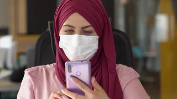 Mulher de negócios muçulmana moderna em hijab e máscara usa um smartphone. Trabalhando no escritório no contexto da pandemia de Covid-19 — Vídeo de Stock