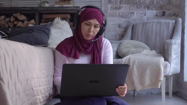 Mulher muçulmana amigável trabalhando remotamente em casa. Bloqueio, bate-papo online, conferência de negócios usando fones de ouvido, microfone e laptop. Trabalho remoto, freelance, educação. Professora de ensino à distância — Vídeo de Stock
