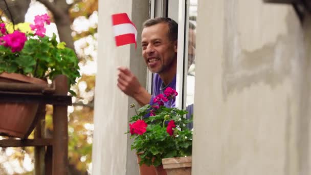 Заблокирован Ковид-19 в Австрии. Австриец на домашнем балконе или окне улыбается и машет флагом Австрии. Самоизоляция в карантине, изоляция, пребывание дома, социальное дистанцирование — стоковое видео