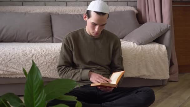 Muselman student läser en bok hemma sitter på golvet. Traditionell religiös skola, distansundervisning, nedstängning. Ung man tonåring studerar — Stockvideo