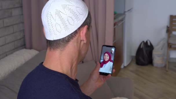 Hombre musulmán asiático hace una videollamada en línea usando un teléfono inteligente y se comunica con una feliz mujer musulmana sonriente en un hiyab. Bloqueo, distancia social durante Covid-19 — Vídeo de stock
