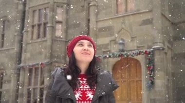 A woman in a red sweater and hat is standing on a European street. The houses are decorated with Christmas lights. Snow is falling. Time of Magic. Holiday season, travel