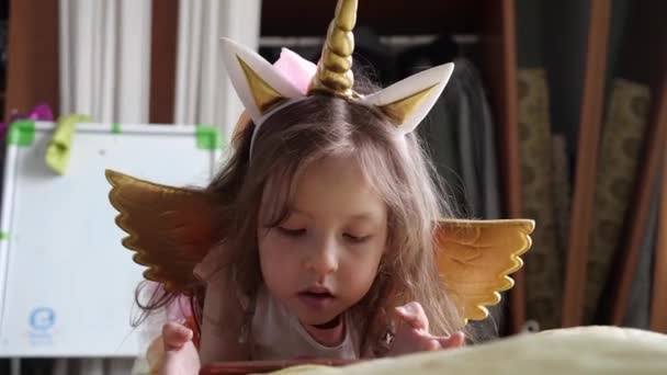 A cute little girl in a unicorn costume with wings is watching a cartoon on her smartphone — Stock Video