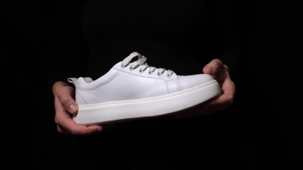 Manos sosteniendo zapatilla de cuero blanco con cordones y mostrando su suela contra fondo negro — Vídeo de stock