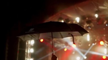 Mutlu insanlar yağmurda şemsiyeler altında harika bir müzik konseri izler. Neşeli hayranlar zıplıyor ve ellerini kaldırıyor. Bir müzik festivalinde çok renkli bir ışık. 