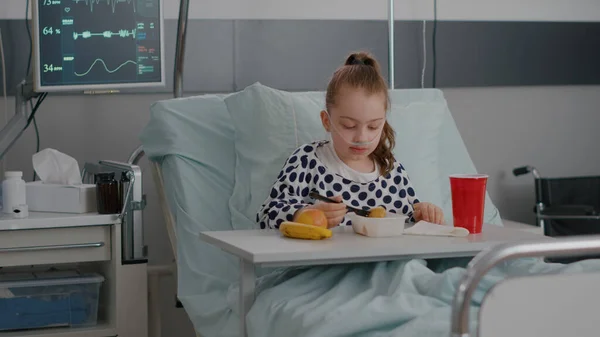 Retrato de un niño pequeño hospitalizado descansando en la cama comiendo comida saludable durante el examen de recuperación —  Fotos de Stock