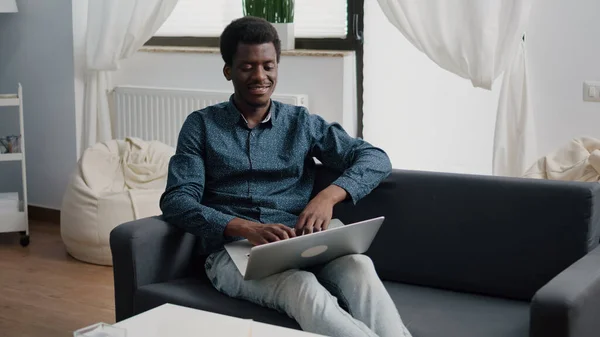 Rilassarsi sul divano, ragazzo afroamericano digitando e lavorando con il computer portatile moderno — Foto Stock