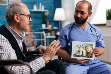 Uzman asist elinde tablet bilgisayar tutarken emekli erkek hasta arkadaşlarıyla selamlaşıyor.