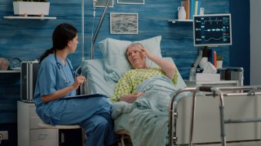 Yatakta hastalıklı kıdemli bir hastayla konuşan tıbbi asistan.