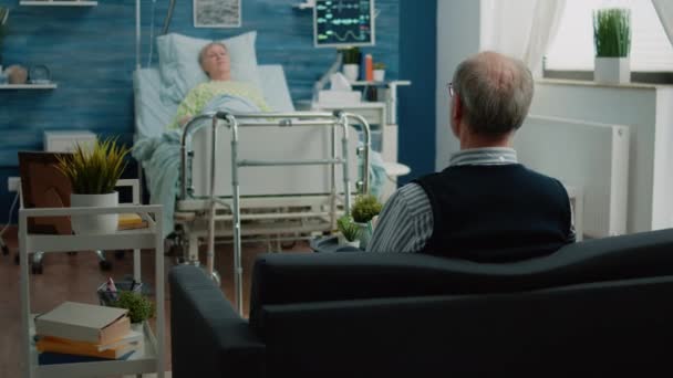 सीनियर मैन सोफे पर बैठे और अस्पताल बिस्तर में बीमार पत्नी से बात कर रहे हैं — स्टॉक वीडियो