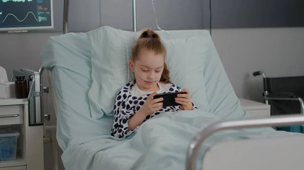 Malade petit enfant reposant au lit jouer à des jeux vidéo en ligne à l'aide d'un smartphone — Photo