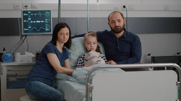 Portrait of sad family looking into camera while holding sick daughter hands