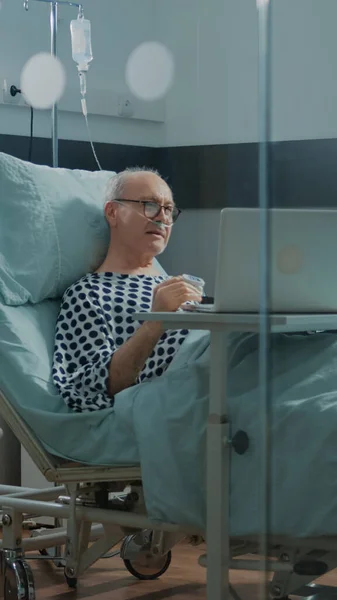 Старый больной пациент с ноутбуком на больничной койке — стоковое фото