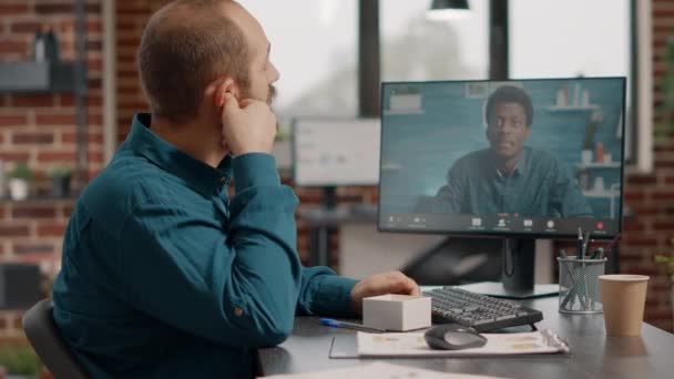 Человек, использующий наушники и видеосвязь на мониторе для общения с коллегой — стоковое видео