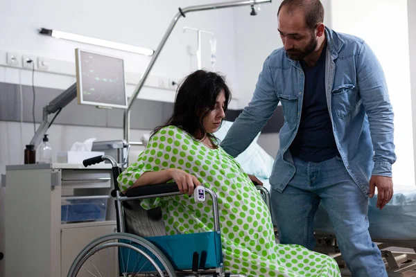Беременная женщина, сидящая в инвалидном кресле, пока муж помогает — стоковое фото