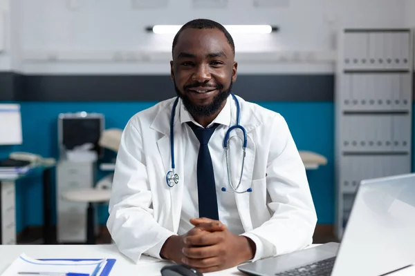 Retrato do sorridente médico especialista afro-americano sentado à mesa no hospital — Fotografia de Stock