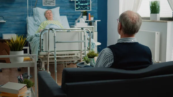 Uomo anziano seduto sul divano e parlare con moglie malata nel letto d'ospedale — Foto Stock