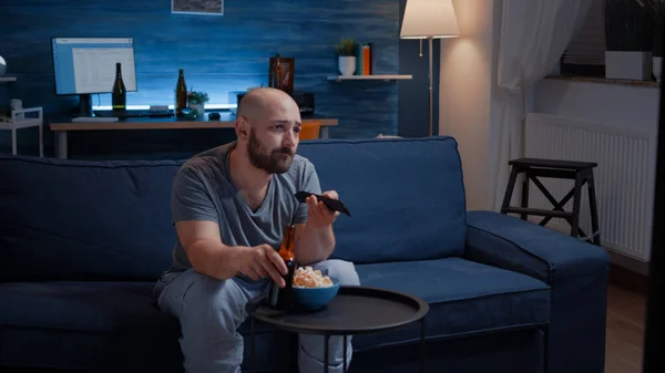 Gelangweilter Mann, der Fernsehsender wechselt, sitzt auf gemütlicher Couch und trinkt Bier — Stockfoto