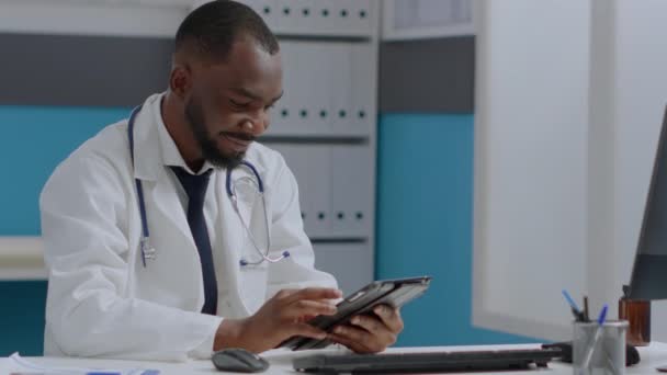 Afroamerikansk terapeut läkare sitter vid skrivbordet på sjukhus kontor håller tablett dator — Stockvideo