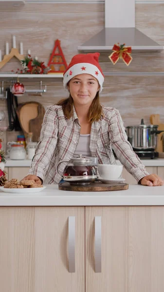 Πορτρέτο του κοριτσιού φορώντας καπέλο Σάντα κοιτάζοντας σε κάμερα στέκεται στο τραπέζι σε Χριστούγεννα διακοσμημένα κουζίνα — Φωτογραφία Αρχείου