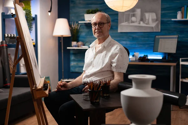 Sanat atölyesindeki atölyede oturan kıdemli sanatçı kameraya bakıyor.