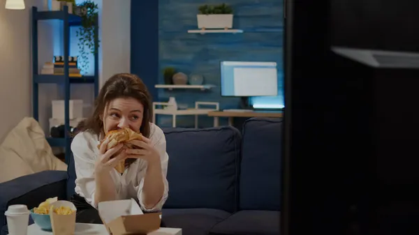 Jovem mulher tomando mordida de hambúrguer sentado no sofá — Fotografia de Stock