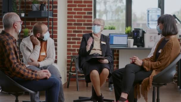 Női terapeuta tanácsot ad az embereknek egy csoportos találkozón, maszkot visel. — Stock videók