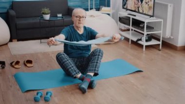Yaşlı yetişkin yoga minderinde oturup direniş bandını çekiyor.