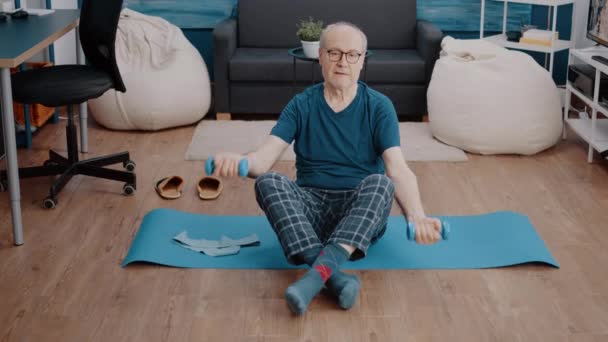 Älterer Mann sitzt auf Yogamatte und stemmt Gewichte, um Muskeln zu trainieren — Stockvideo