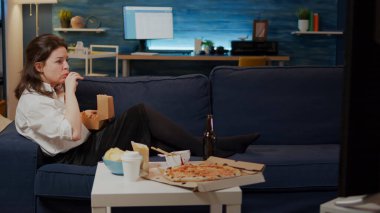 Genç kadın hamburger yerken kanepede uzanıyor.