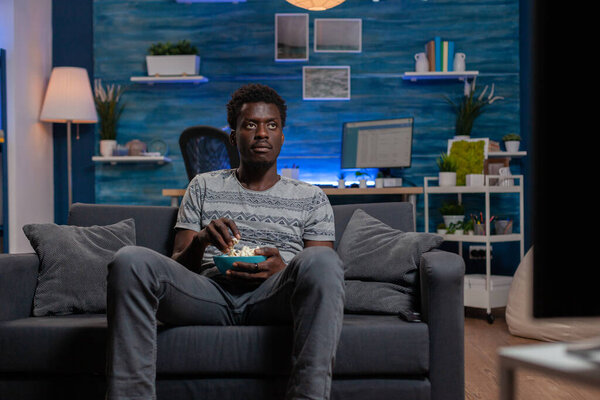 African american guy resting on sofa in living room holding popcorn bowl
