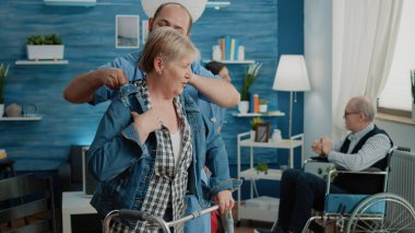 Hemşire yardım ederken engelli kadın ceket giyiyor.