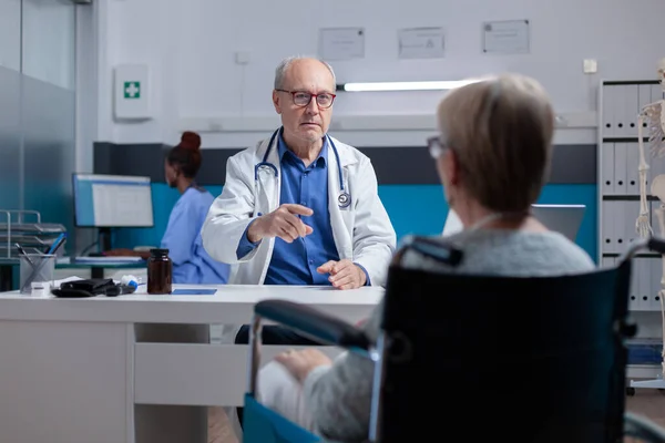 Doctor attending medical consultation with woman sitting in wheelchair at clinic — Stock Photo, Image