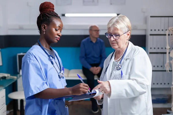 의사가 캐비닛에서 클립보드를 들고 있는 동안 아프리카 계 미국인 간호사가 수표에 서명을 하는 모습 — 스톡 사진