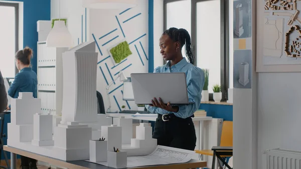 Mulher arquiteta segurando laptop para analisar modelo de edifício no escritório de arquitetura. — Fotografia de Stock