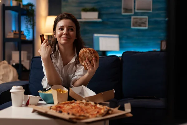 Vrouw die lekkere afhaalmaaltijden fastfood hamburger eet terwijl ze geniet van een koud biertje voor de televisie — Stockfoto