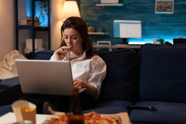Жінка їсть картопляний чіп перекус, що працює на ноутбуці під час перегляду серіалів по телевізору — стокове фото