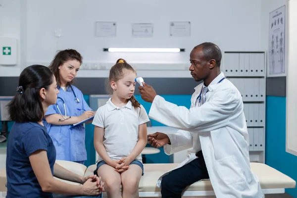 Médico pediatra afroamericano que coloca termómetro infrarrojo médico en la frente de la niña —  Fotos de Stock