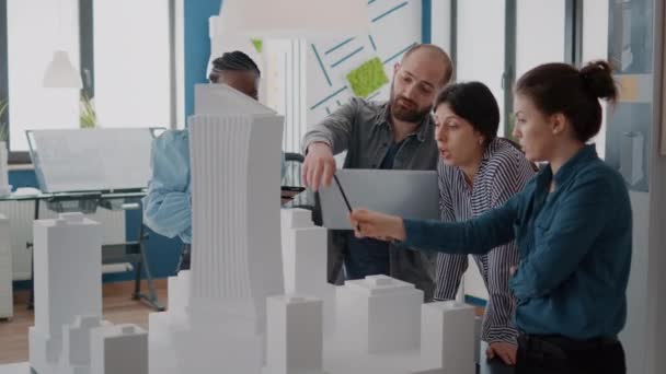 Мультиэтнические люди, работающие с планшетом и ноутбуком для анализа модели здания на столе — стоковое видео