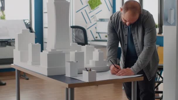 Ingeniero hombre analizando el modelo de edificio para diseñar planos planos en la mesa — Vídeo de stock