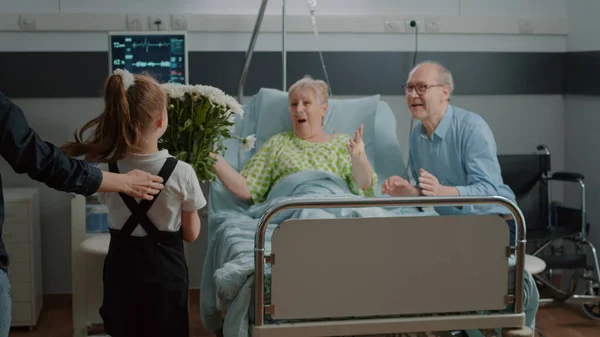 Klein meisje rennen om te knuffelen en bloemen te geven aan oudere patiënt in ziekenhuisafdeling bed — Stockfoto