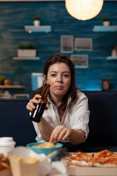 Porträt einer Frau, die in die Kamera blickt, eine Bierflasche hält und Kartoffelchips aus der Schüssel isst — Stockfoto