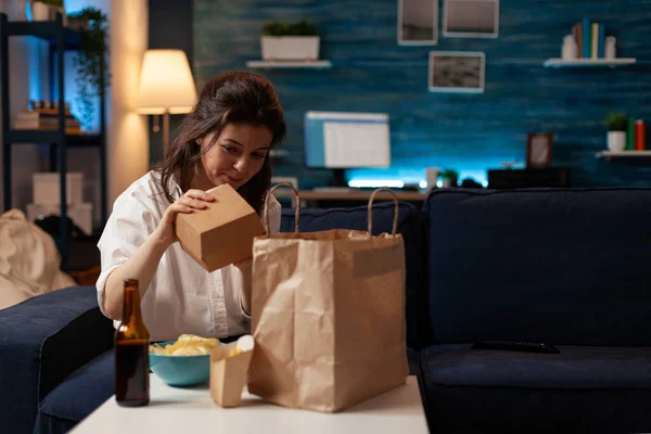 沙发上的女人从纸袋中取出汉堡包，坐在桌前的沙发上 — 图库照片