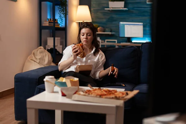 Mulher cansada comendo saboroso hambúrguer takeaway segurando garrafa de cerveja relaxante no sofá — Fotografia de Stock