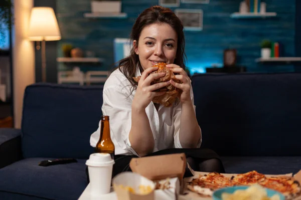 Усміхнена жінка тримає смачний бургер в руках, дивлячись на телевізійну комедію ситкому, що стоїть на дивані — стокове фото