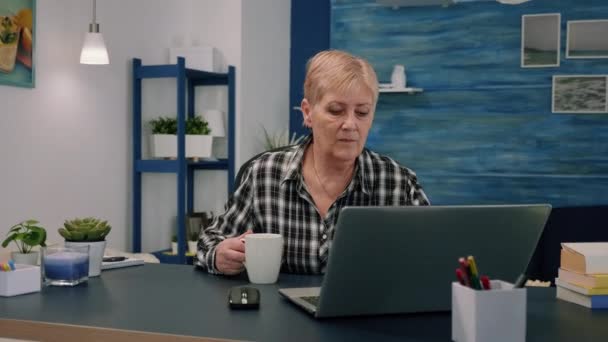 Παλιά επιχειρηματίας κάθεται στο χώρο εργασίας δακτυλογράφηση στο φορητό υπολογιστή, πίνοντας καφέ — Αρχείο Βίντεο