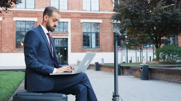 Бизнесмен в дипломатическом костюме сидит на скамейке, печатая стратегию компании на ноутбуке — стоковое фото
