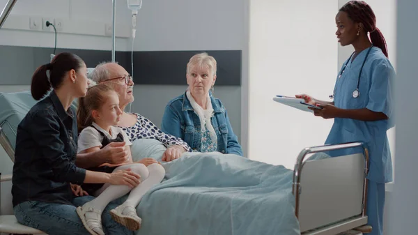 Сім'я, яка відвідує пенсіонера в лікарняному відділенні і розмовляє з медсестрою — стокове фото