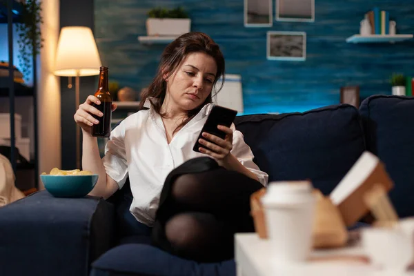 İşten sonra kanepede dinlenen kadın elinde bira şişesi olan akıllı telefonuyla sosyal medyada geziniyor. — Stok fotoğraf