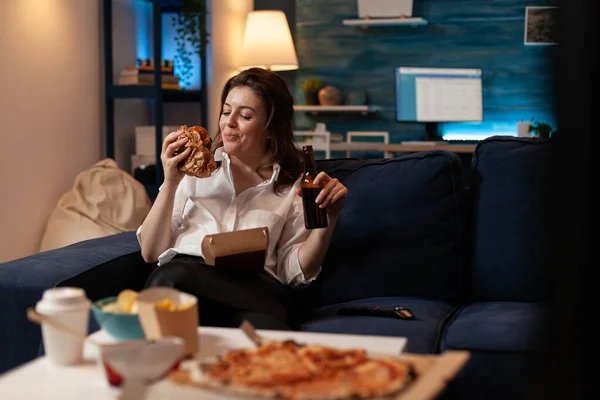 Smiling woman sitting on couch looking at tasty takeaway burger holding beer bottle in front of table witht pizza